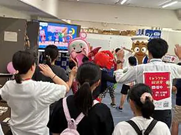 かまぼこやちくわのキャラクターの着ぐるみと一緒に、子どもたちが画面を見ながら体操をしている。